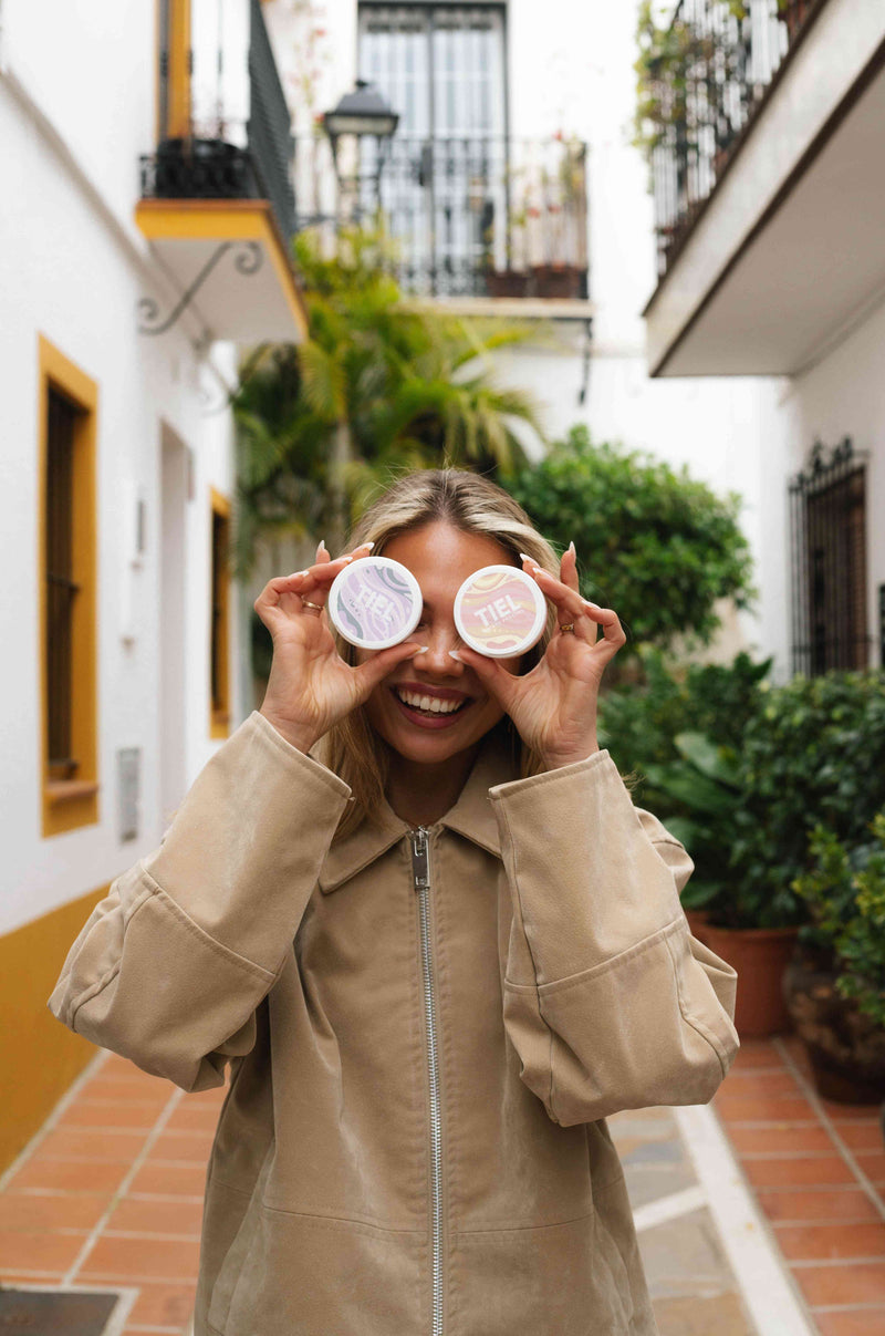 Glad kvinna håller två TIEL energy pouches dosor framför ögonen – viol lakrits och peach mango nikotinfritt snus med koffein